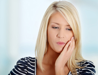 Head-and-shoulders photo of a woman with shoulder-length blonde hair. She is wearing a navy and white top and is holding the left side of her upper cheek; for information on San Antonio emergency dentistry at My Smile Artist.